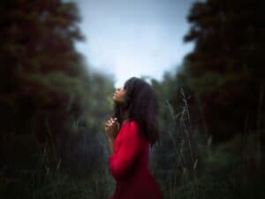 Exploring the Transformational Power of Surrender Lady standing before a forest in a red dress, looking towards the sky with hands clasped and eyes closed in prayer