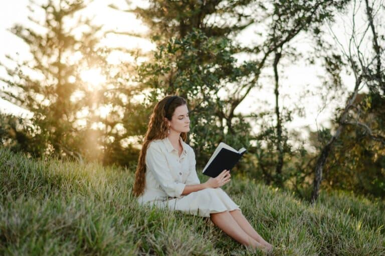 Home Lady in cream dress reading the Bible while sitting on the grass with trees behind her