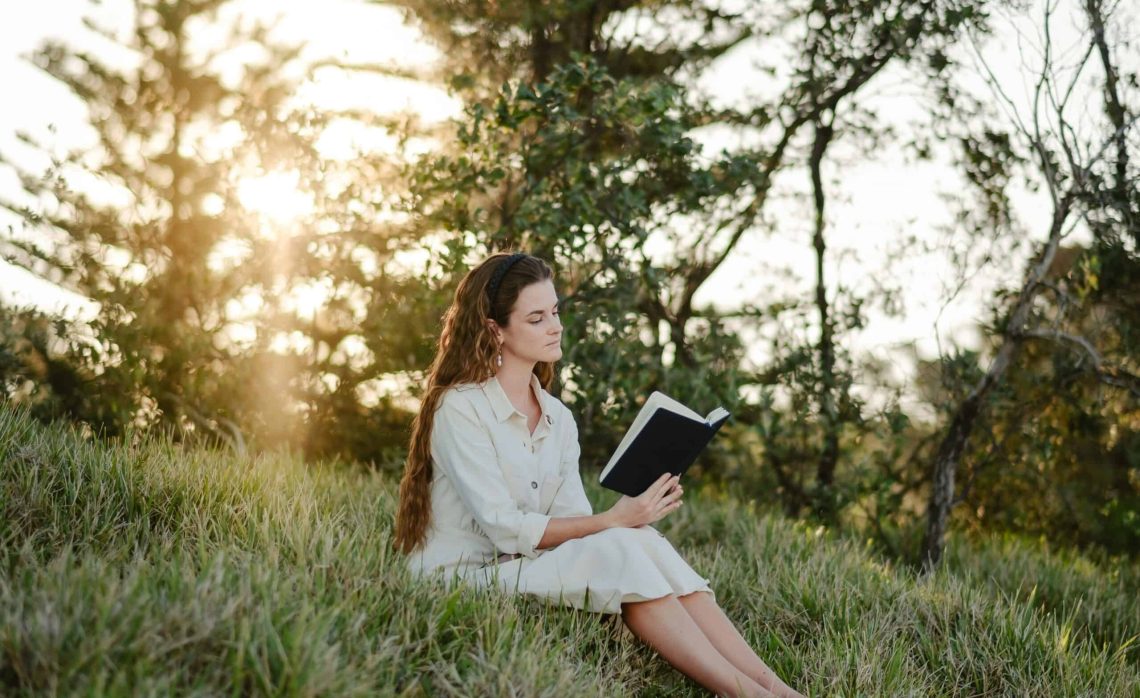Lady in cream dress reading the Bible while sitting on the grass with trees behind her